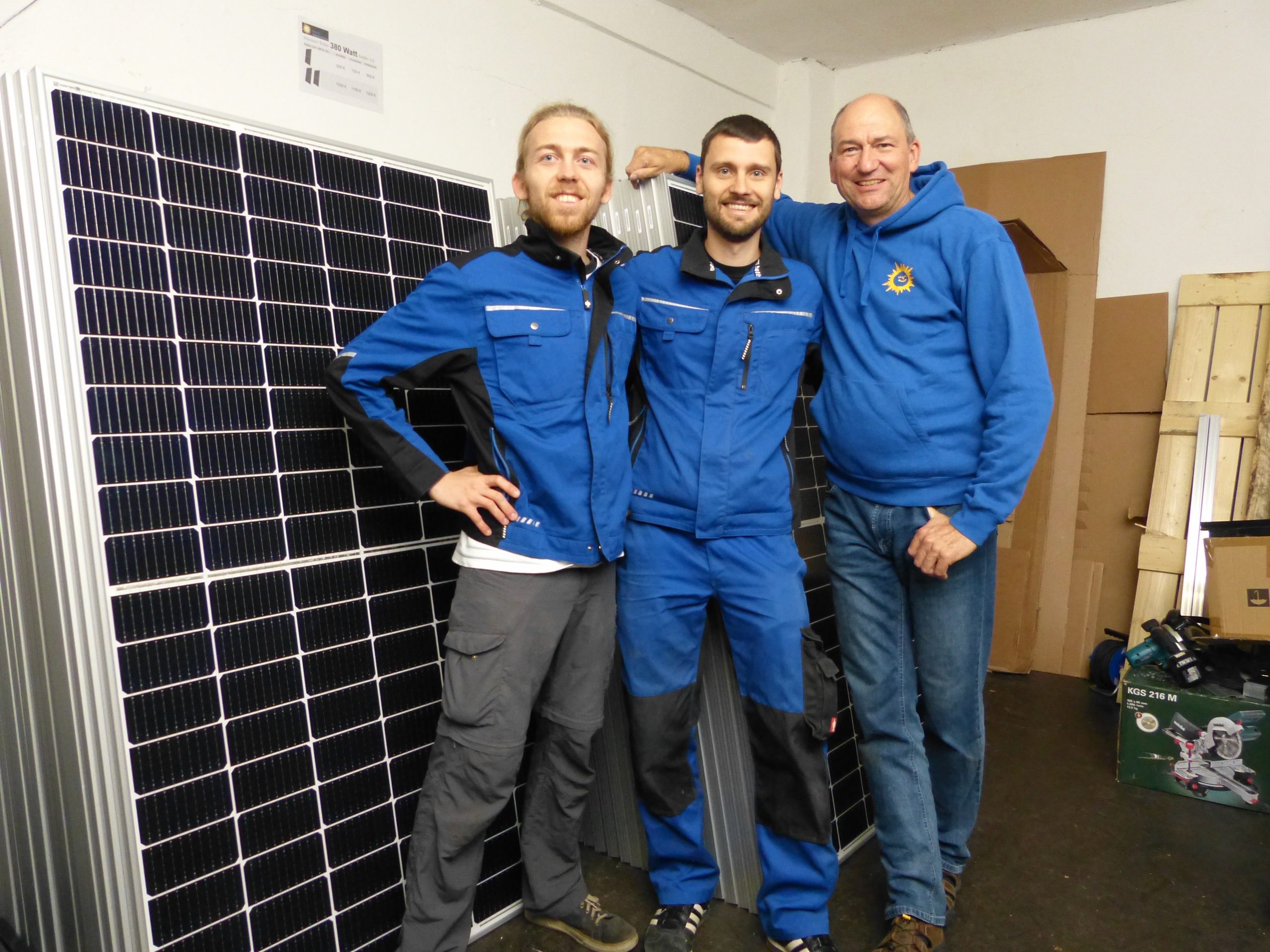 Drei blau gekleidete, männlich und weiß gelesene Personen stehen vor Solarmodulen in einem Lagerraum und lächeln